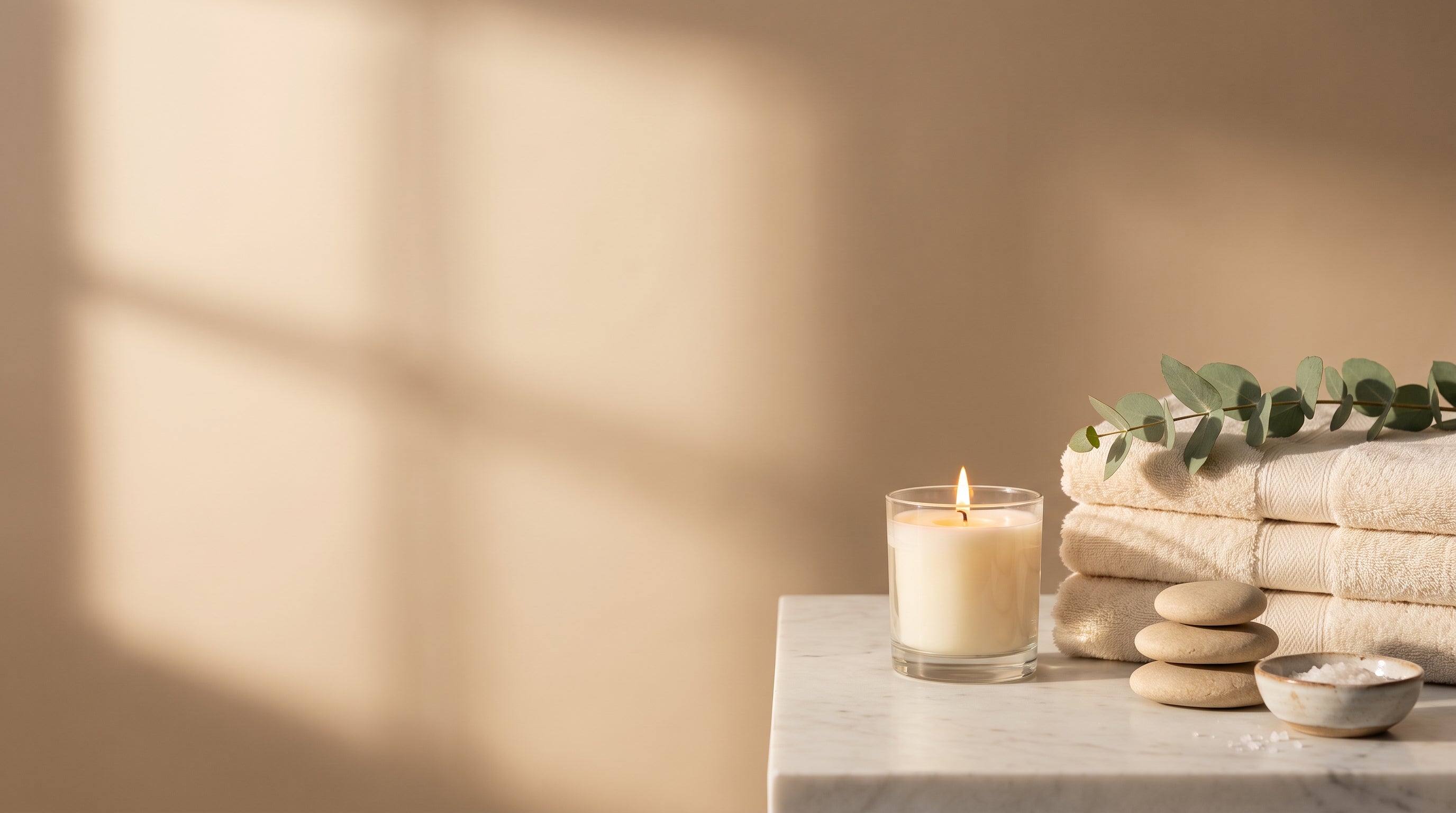 Homepage hero - warm spa still life with candle, towels, eucalyptus and massage stones on marble, copy space left